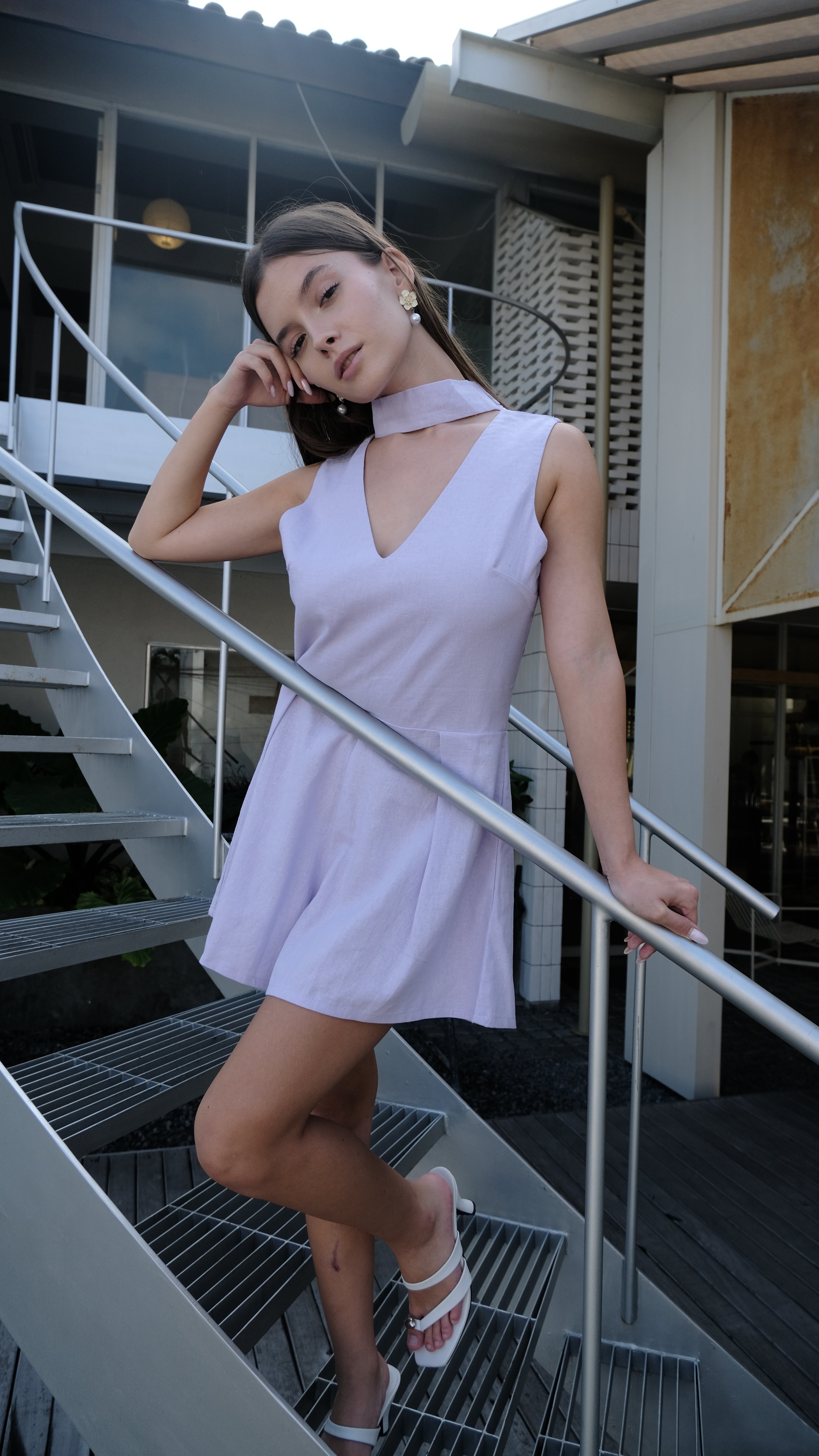 A woman wearing a light lavender sleeveless mini dress with a keyhole cutout, posing dynamically on a modern metal spiral staircase.